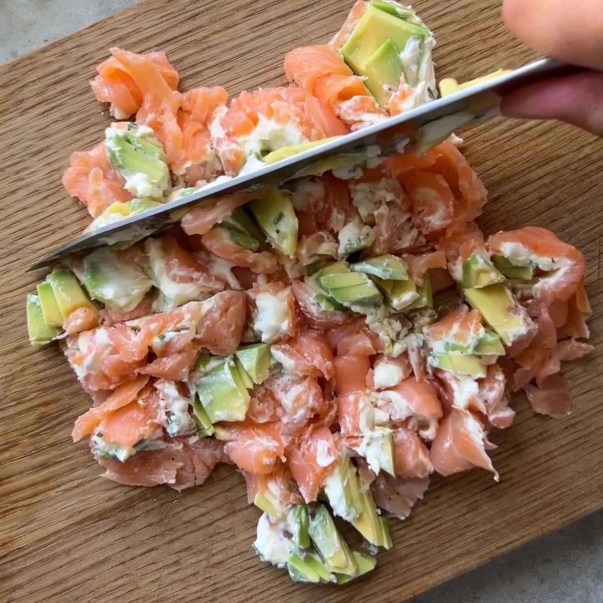 A knife slices through a vibrant mixture of smoked salmon, avocado, and cream cheese on a wooden cutting board.