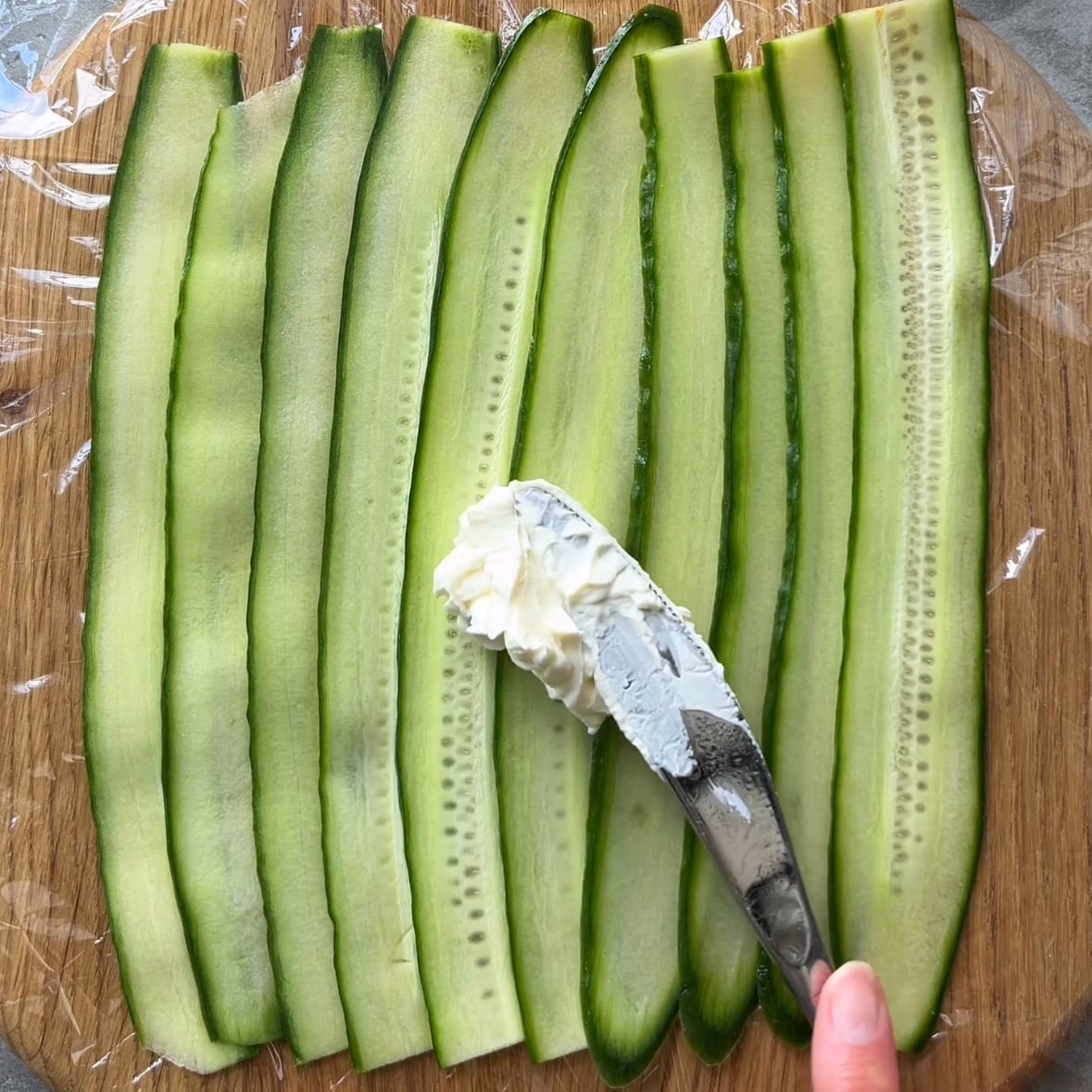 Thin cucumber slices are arranged on a wooden board lined with plastic wrap, and a knife is spreading cream cheese onto them.