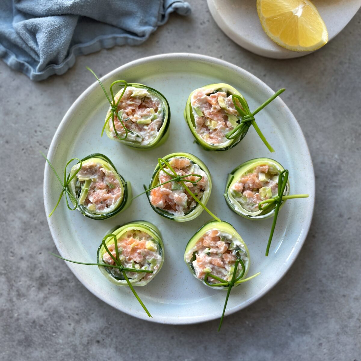 A plate with seven cucumber bites filled with a salmon mixture, garnished with chives, sits on a gray surface beside a lemon wedge and a folded blue tea towel.