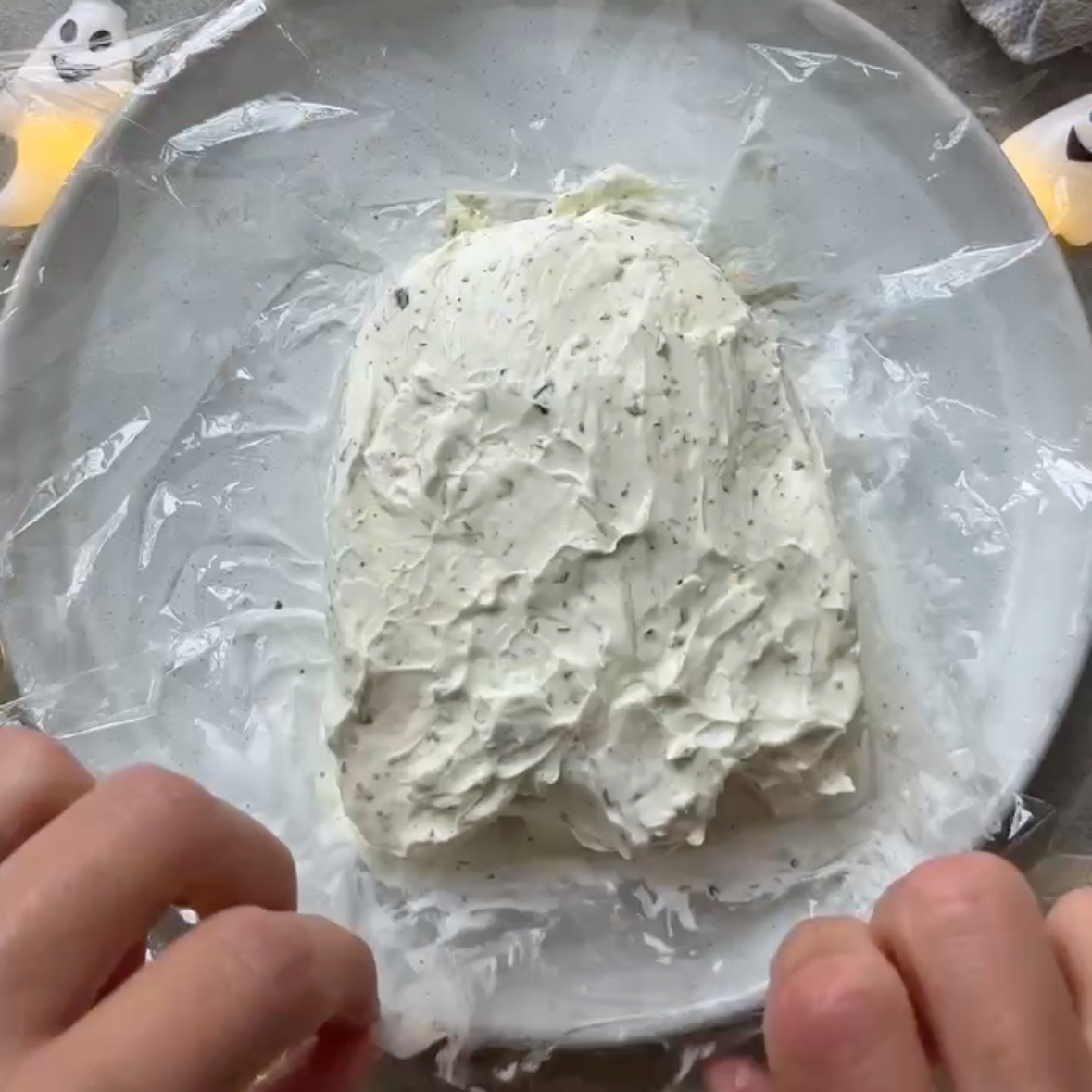 Hands are wrapping up a ghost shaped cheese ball into cling film.