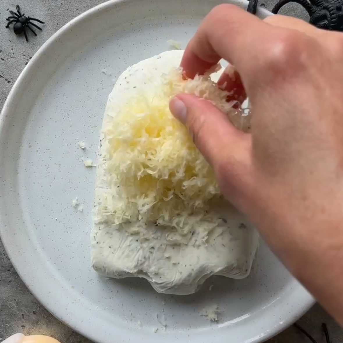 A hand topped grated cheddar cheese on top of a ghost shaped cheese ball.
