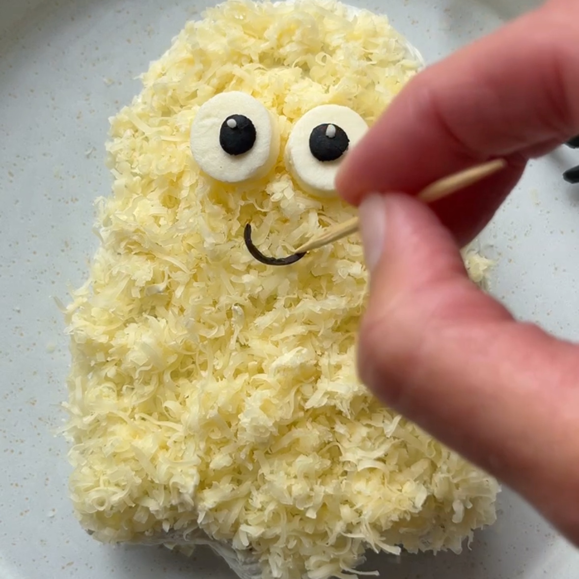 A hand uses a toothpick to place an olive smile on to a ghost cheese ball.