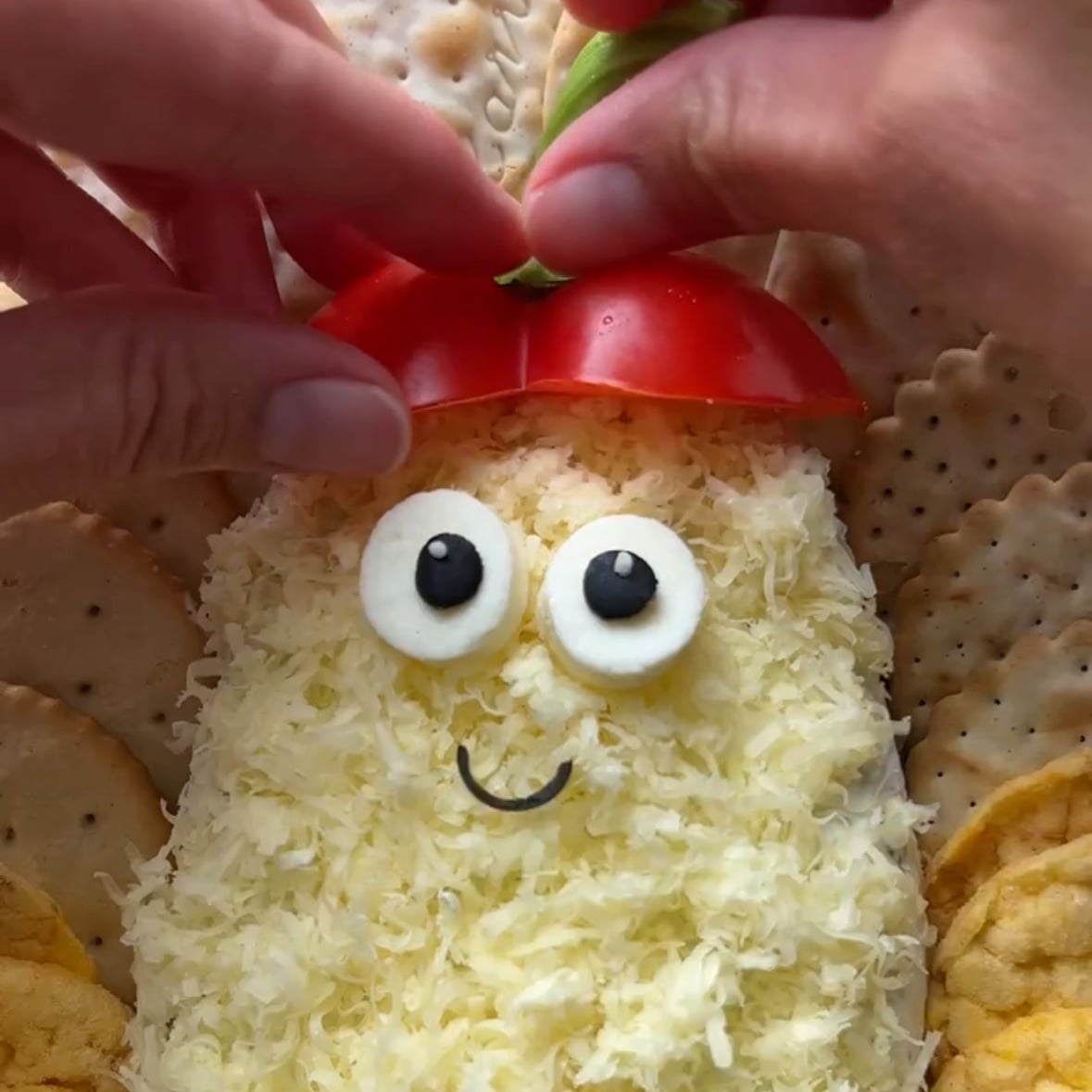 Hands arrange a red pepper hat on top of a ghost cheese ball as the final step before serving.