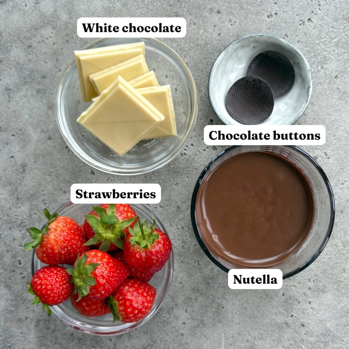 Bowls containing white chocolate squares, chocolate buttons, fresh strawberries, and a glass of Nutella.