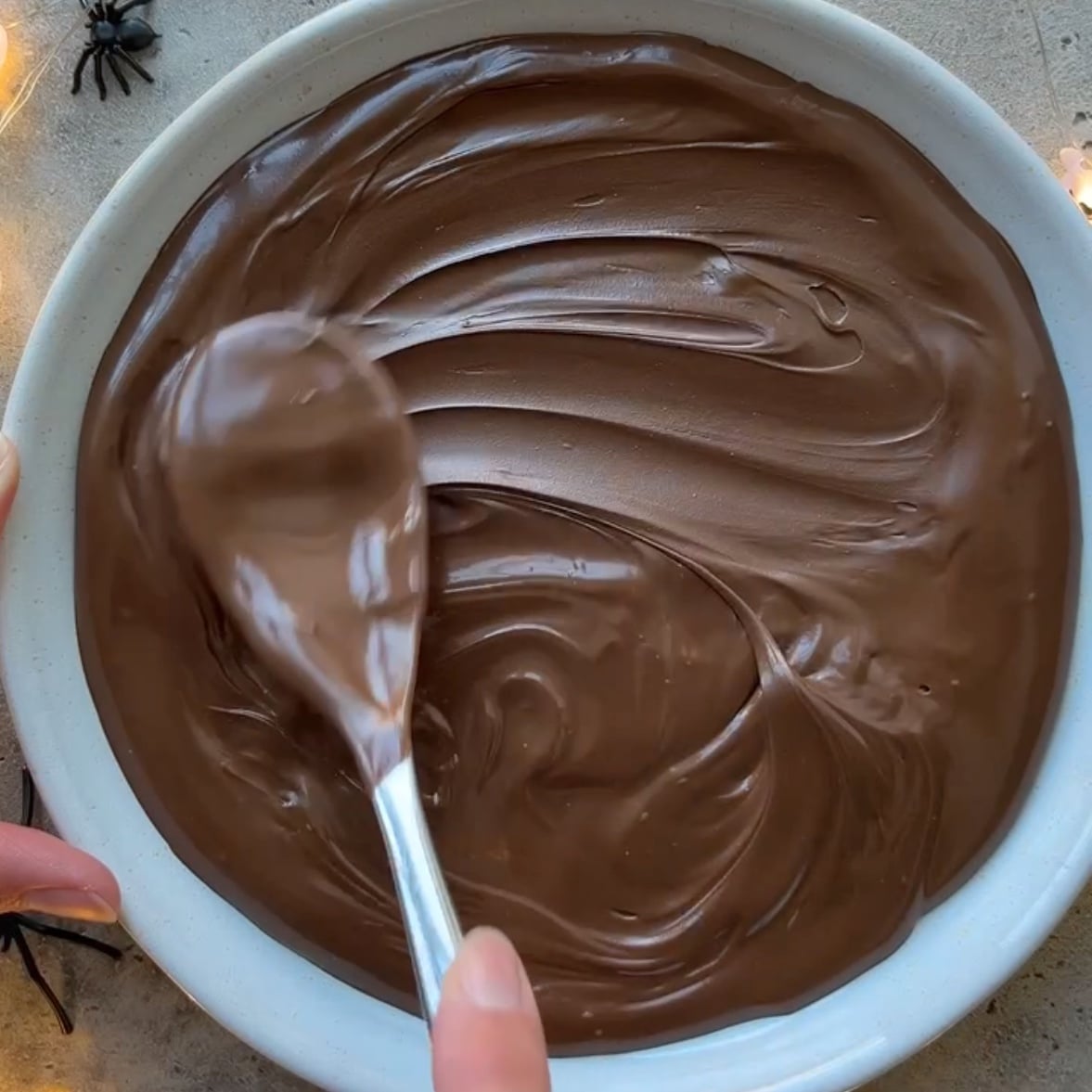 A hand uses a spoon to spread Nutella on a white platter, preparing a Halloween Dessert Dip. Decorative plastic spiders are visible on the table nearby.