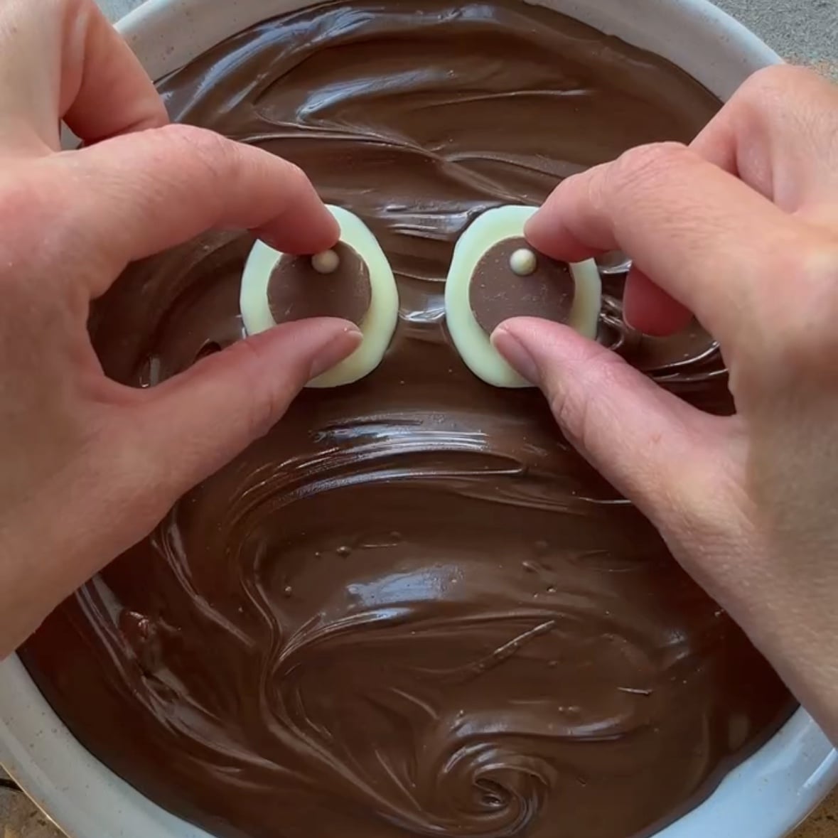 Two hands place chocolate eyes on top of a dish filled with swirled Nutella.