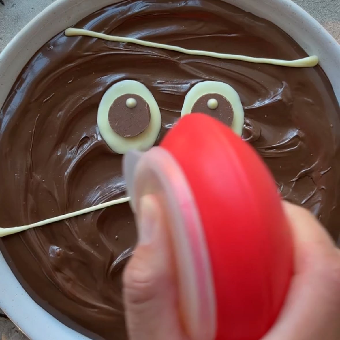A hand holds a red piping tool over a dish of Nutella decorated with a cartoon face made from white and brown chocolate.