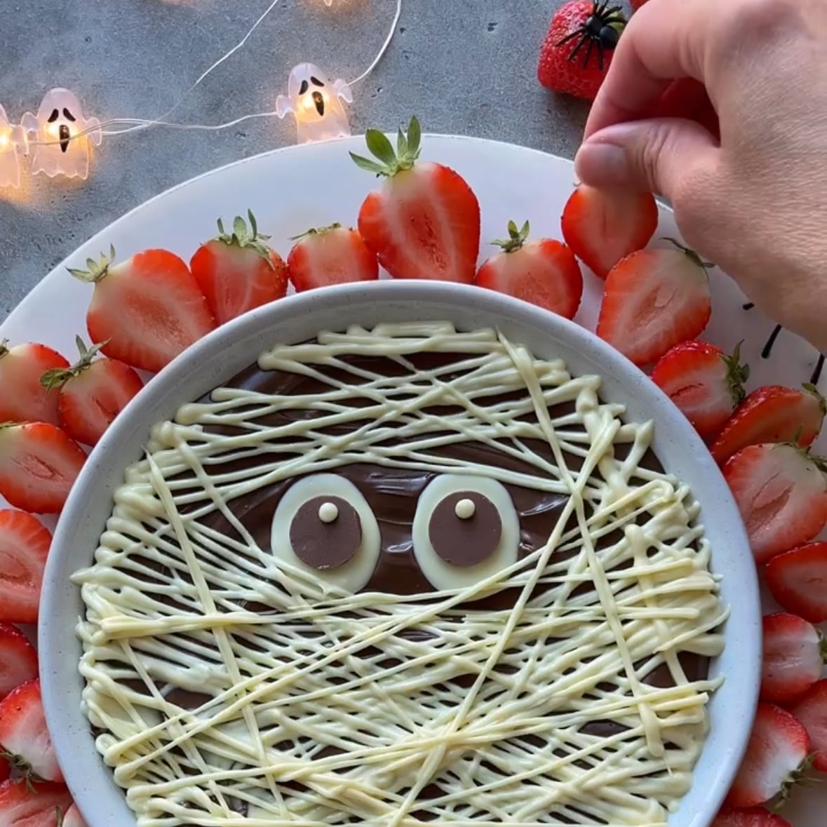 A round Halloween Dessert Dip is decorated like a mummy with white chocolate lines and two candy eyes, surrounded by sliced strawberries on a white plate. A hand is placing a strawberry slice. Ghost lights decorate the scene.