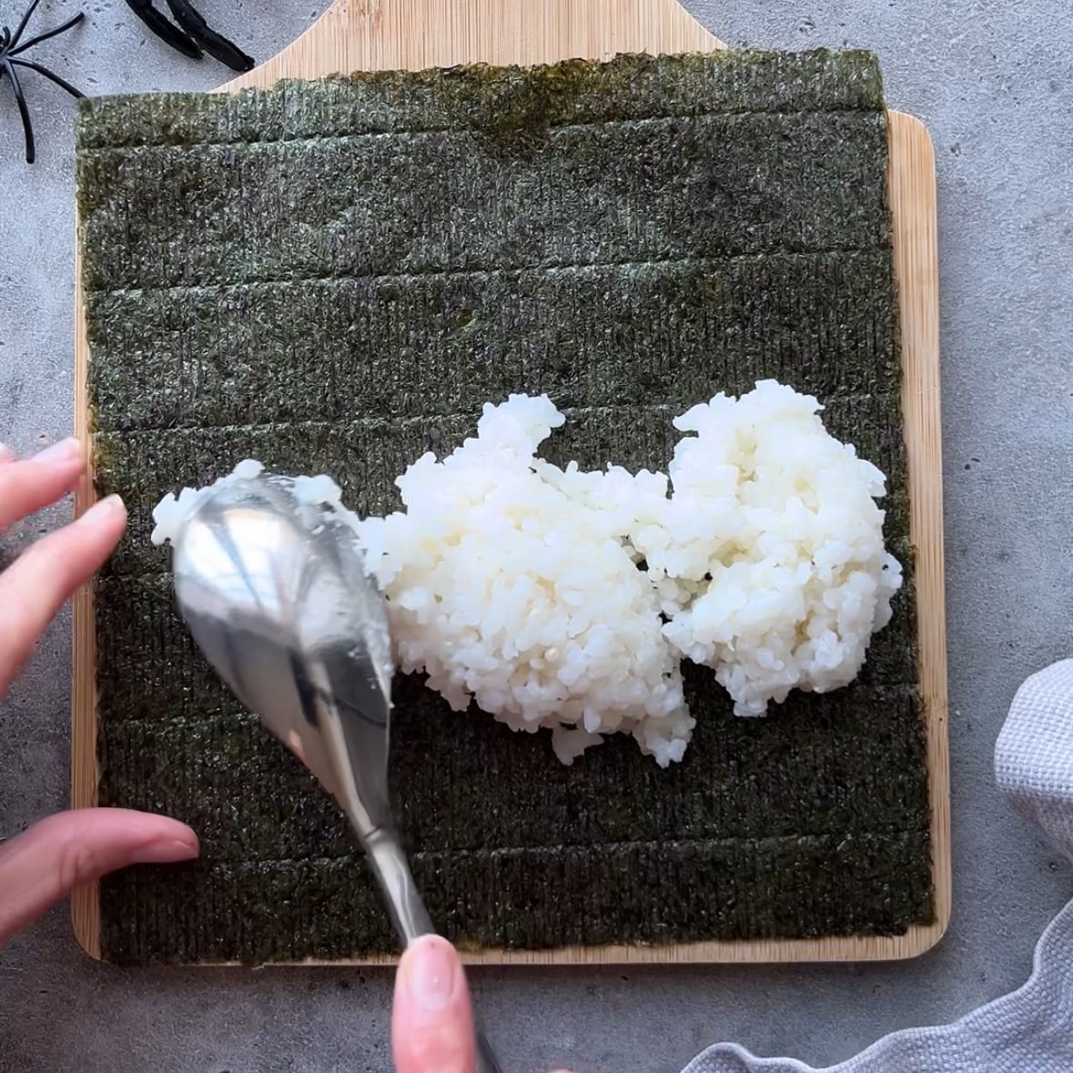 A hand spreads white rice onto a sheet of seaweed using a spoon, all placed on a wooden board with a gray background.