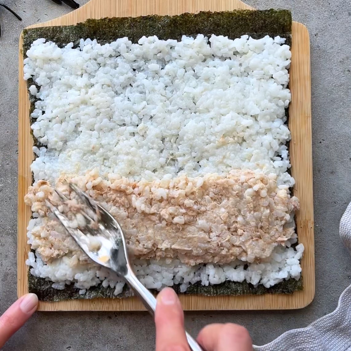 A hand spreads a tuna mixture over sushi rice on a sheet of nori, placed on a wooden board.