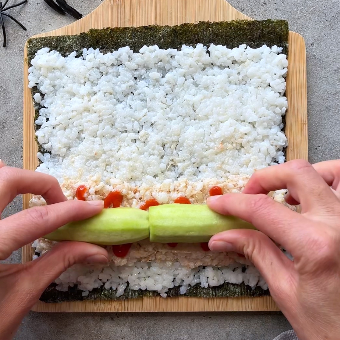 Hands placing cucumber slices on rice and seaweed, with a line of sriracha sauce visible.