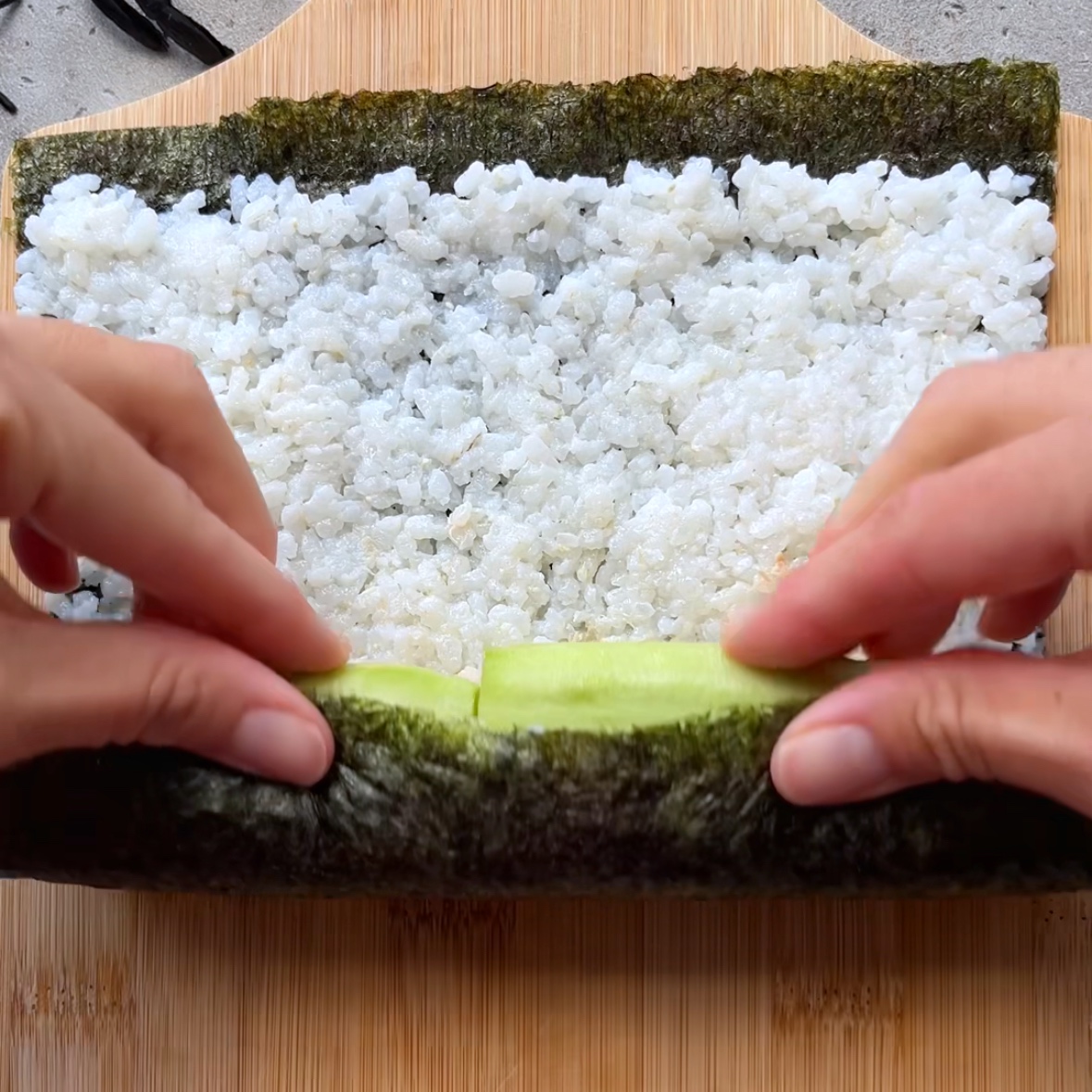 Two hands roll Halloween Sushi with rice and cucumber slices on a sheet of nori, placed on a chopping board.