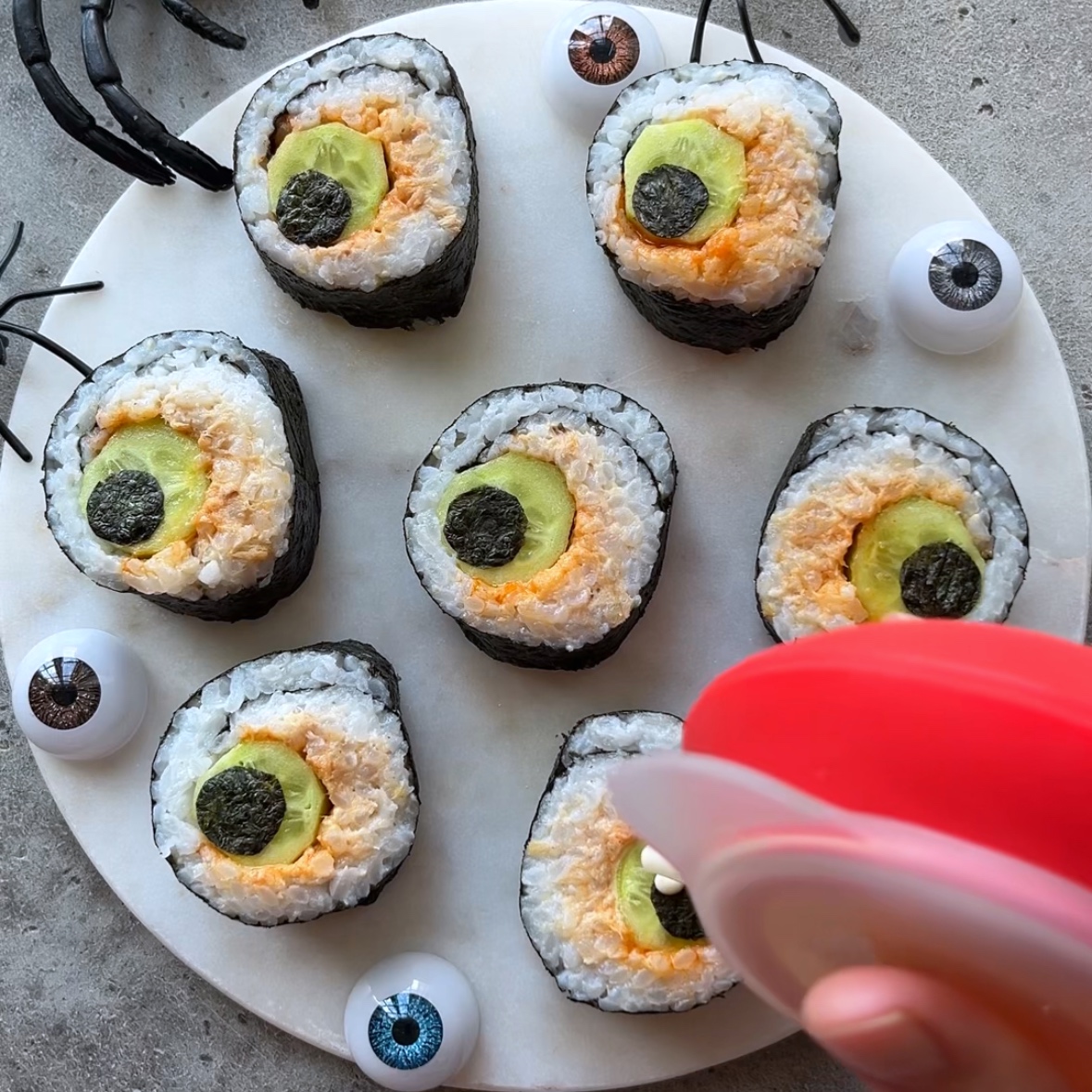 Round sushi rolls arranged on a white plate, designed to look like eyeballs for Halloween Sushi, with fake plastic eyeballs and spider decorations nearby. A hand is squeezing mayo onto one of the sushi rolls.