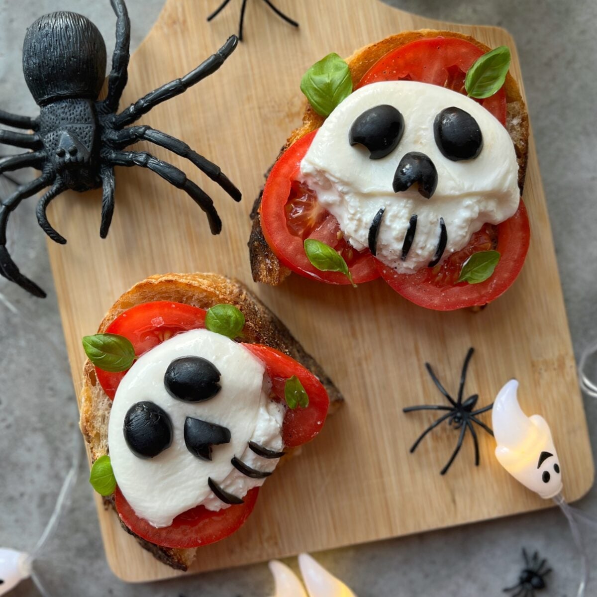 Two slices of toast topped with tomato, mozzarella eyeballs, and olives arranged to look like skull faces, placed on a cutting board with plastic spiders and ghost decorations nearby.