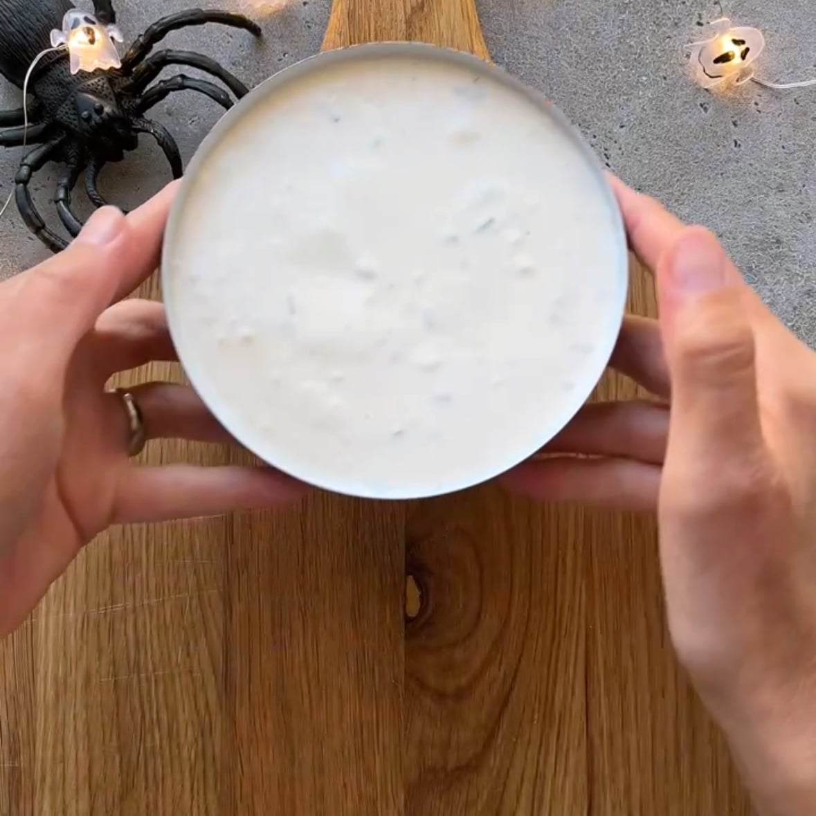 Two hands hold a bowl of white dip on a wooden surface, surrounded by Halloween decorations including a fake spider and skull string lights in the background.
