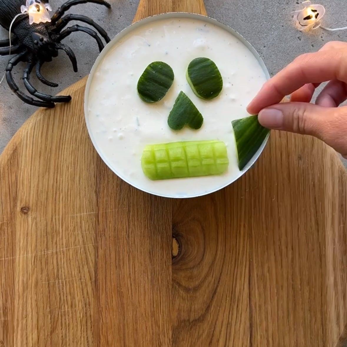 A bowl of white dip is decorated with cucumber pieces to look like a skull face. A hand adds a cucumber piece, while a plastic spider and string lights are nearby on a wooden board.