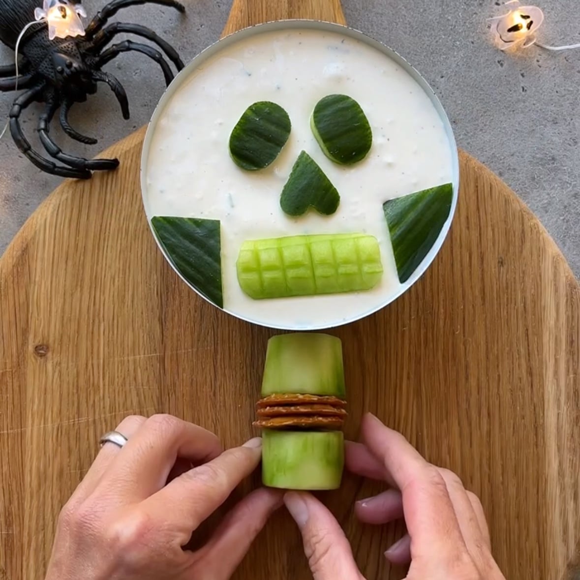 A round dip topped with cucumber slices arranged like a skull, with a cucumber and pretzel spine sits on a wooden board as part of a creative Halloween Veggie Tray. Hands are assembling the spine, while a plastic spider and festive lights are nearby to complete the spooky scene.