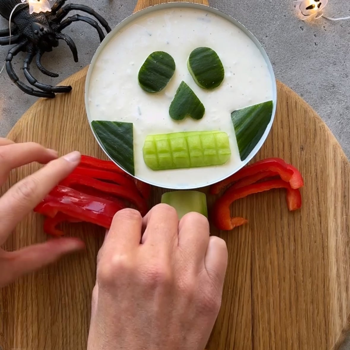 A person arranges red bell pepper slices as ribs on a Halloween Veggie Tray shaped like a skeleton face, complete with cucumber features.