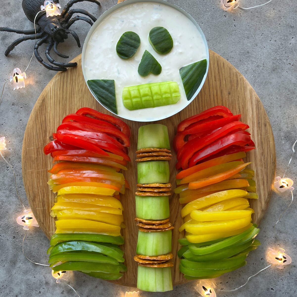 Vegetable platter arranged to resemble a skeleton with bell peppers, cucumbers, crackers, and a dip shaped as a skull with festive decor surrounding the board.