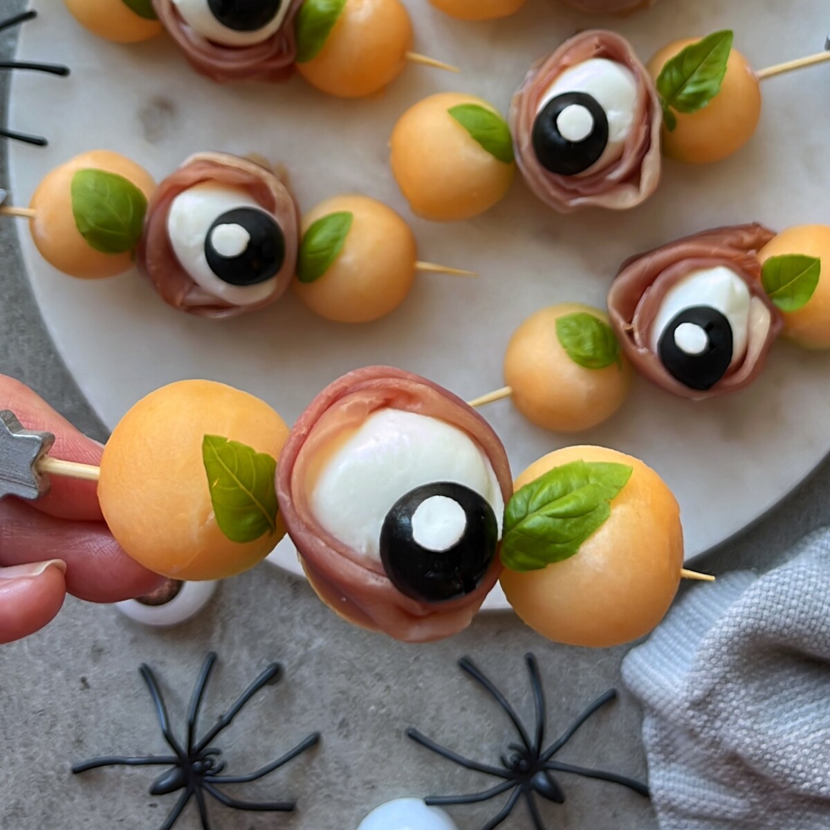 Skewers with melon balls, mozzarella, prosciutto, black olives, and basil arranged to look like eyeballs make fun bites—perfect for party food recipes. Presented on a round white marble tray with decorative plastic spiders nearby.