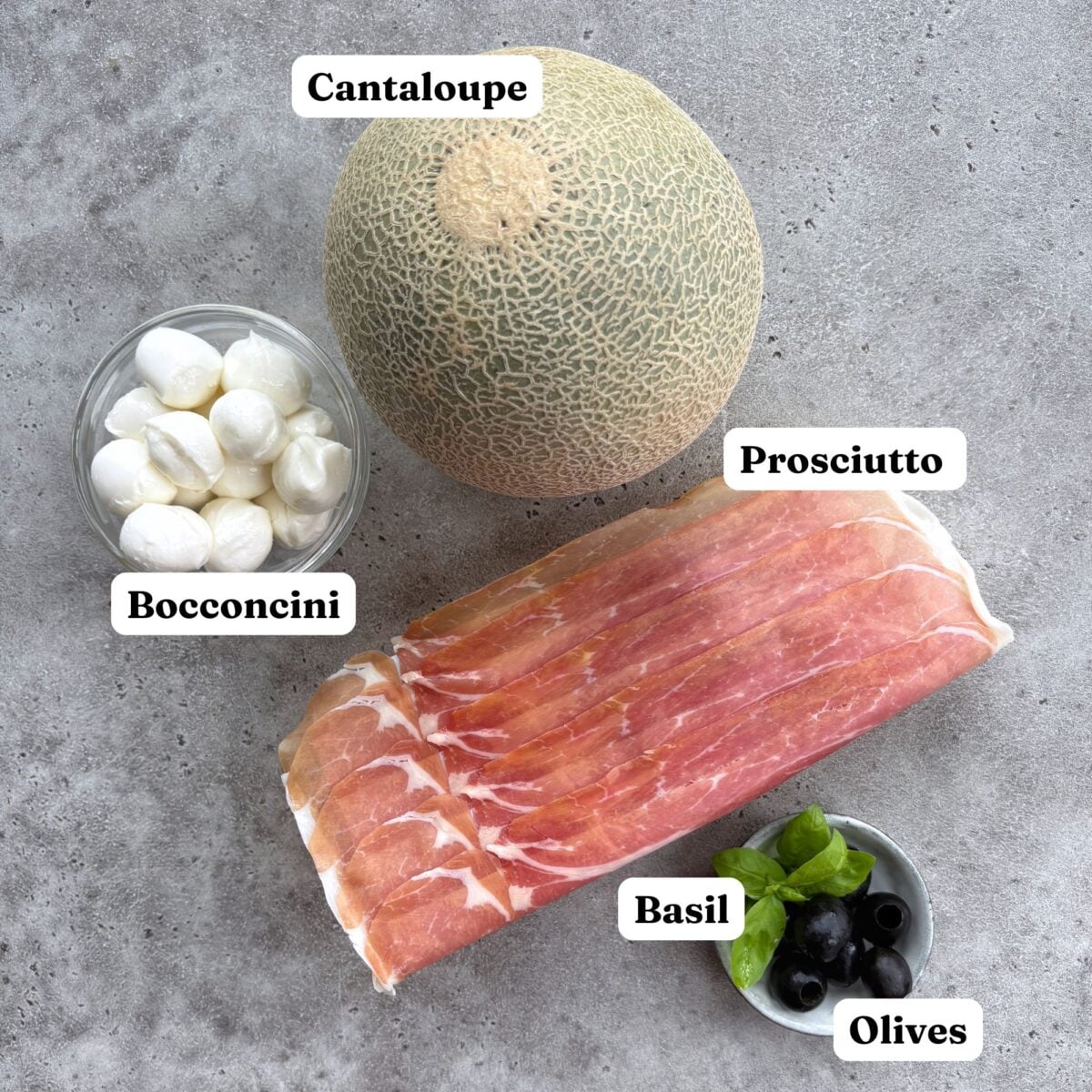 A cantaloupe, a bowl of mini mozzarella balls, a stack of prosciutto, and a small dish of basil and olives.