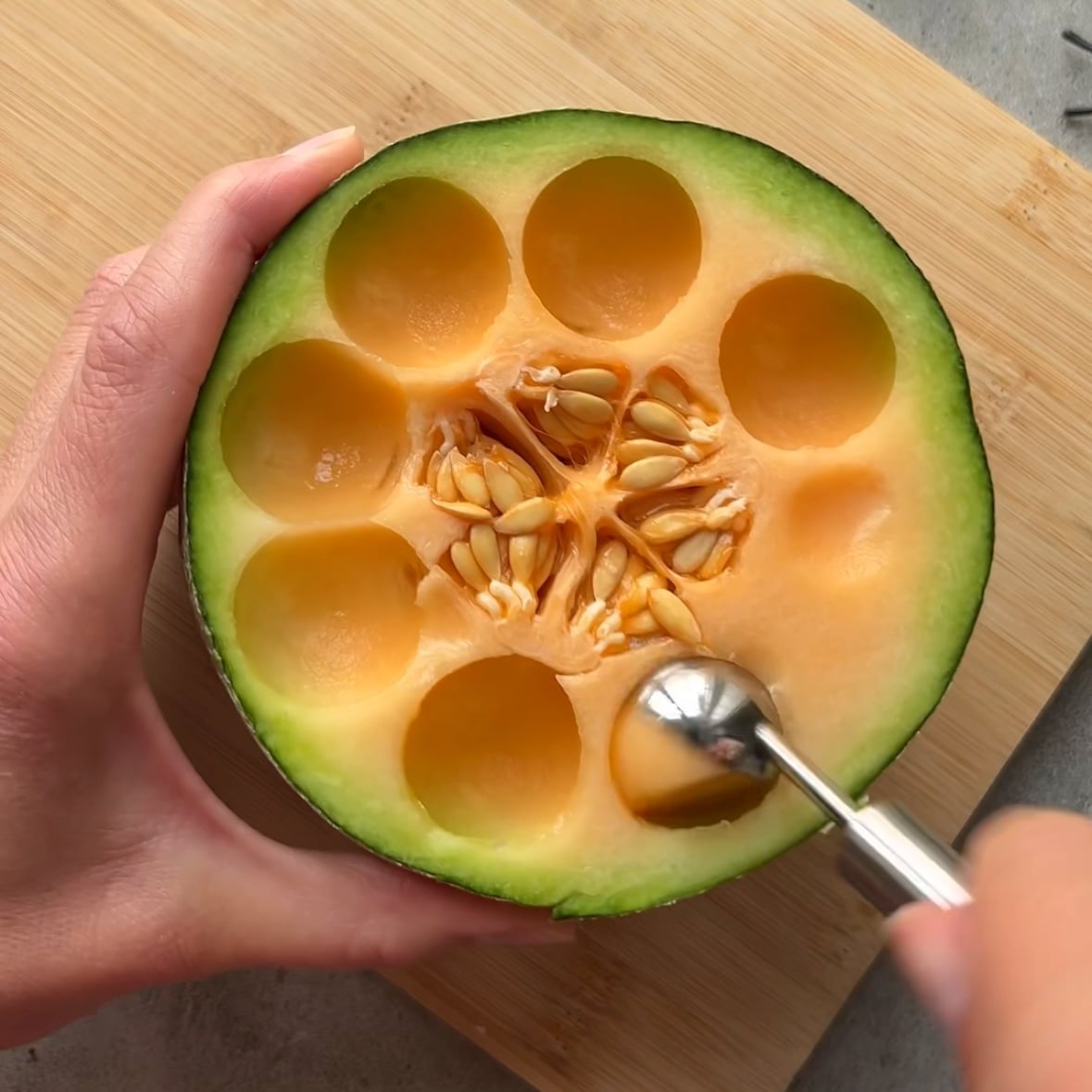 A halved cantaloupe with seeds exposed, and a hand uses a melon baller to scoop out round pieces on a wooden cutting board.