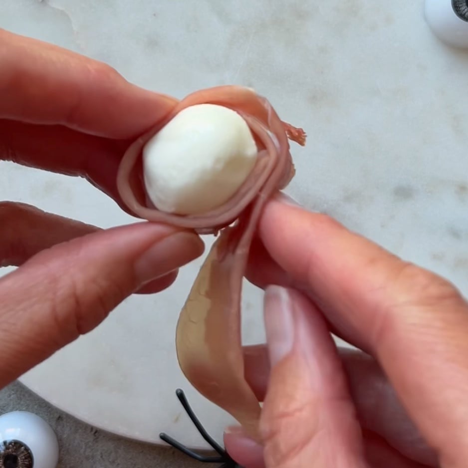 A person wraps a slice of prosciutto around a small ball of mozzarella cheese.