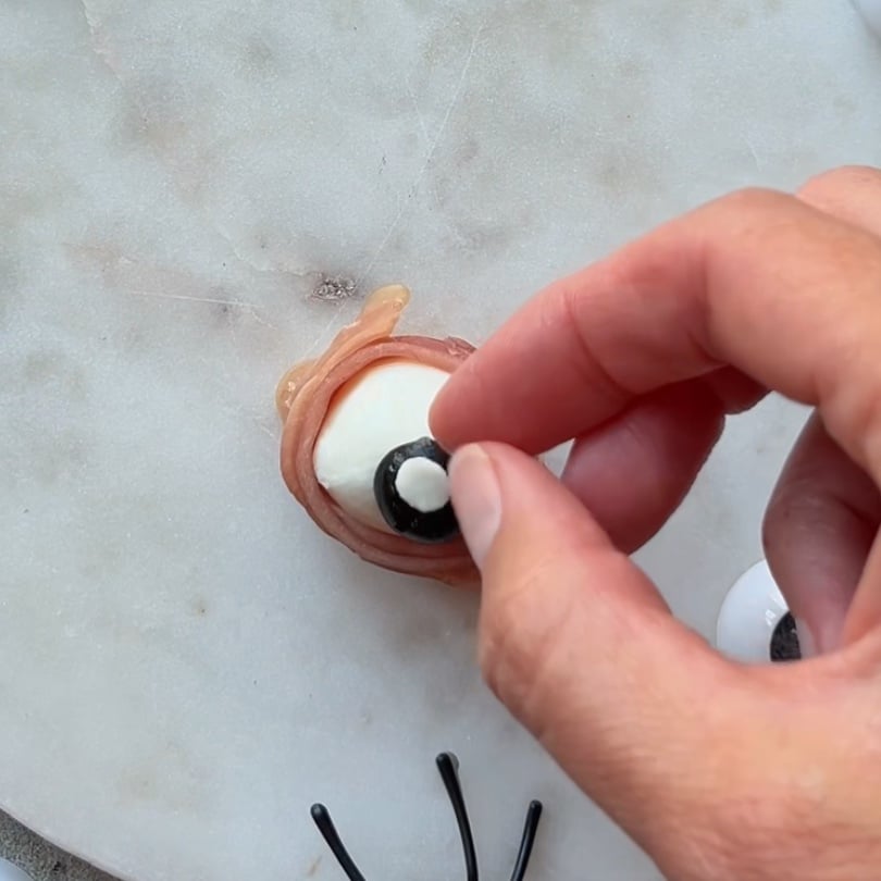 A hand places a black olive slice onto a mozzarella ball wrapped in prosciutto, creating Mozzarella Eyeballs that resemble realistic eyes on a white surface.