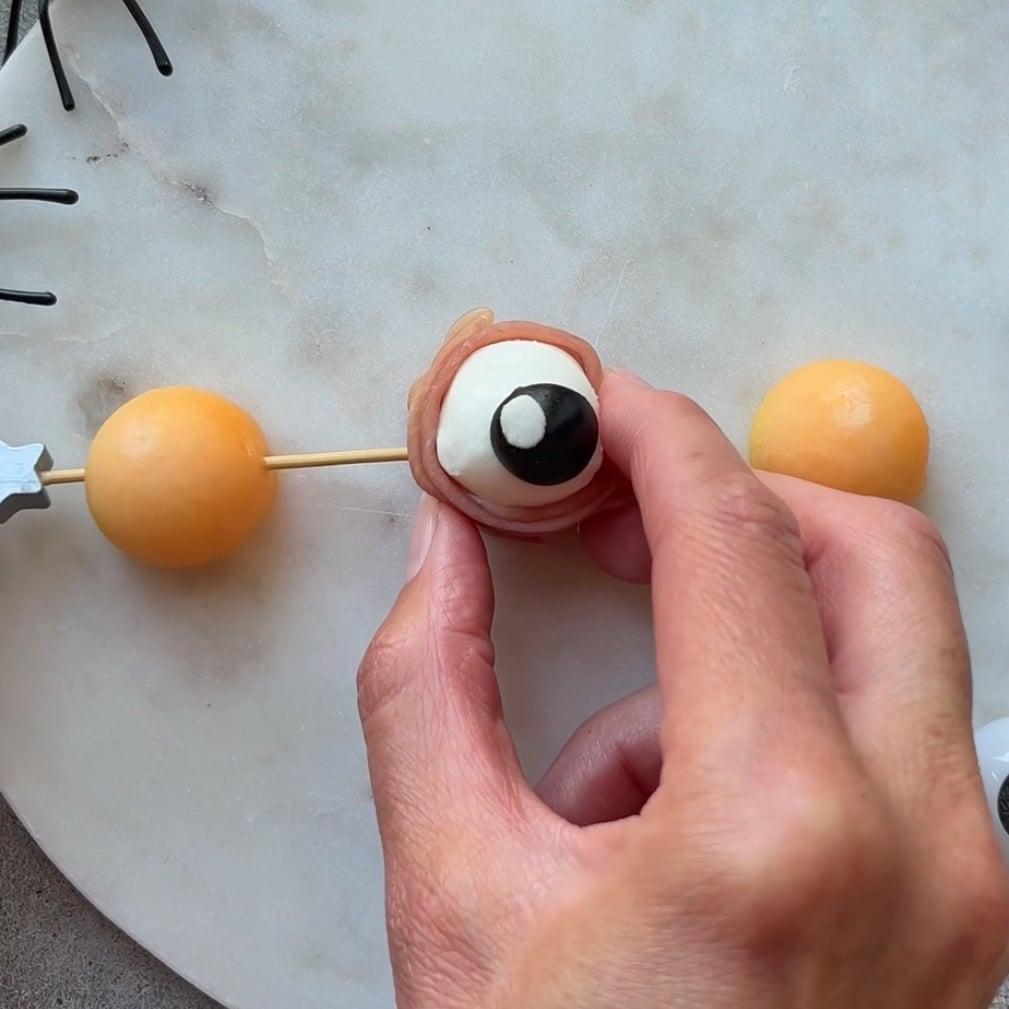 A hand holds a skewer with melon balls and a mozzarella eyeball wrapped in prosciutto on a marble surface.