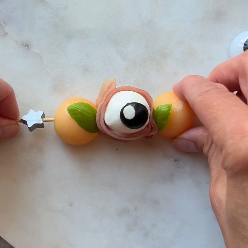 Hands assembling a skewer with two melon balls, basil leaves, prosciutto, and a Mozzarella Eyeball decorated to look like an eyeball, on a marble surface.
