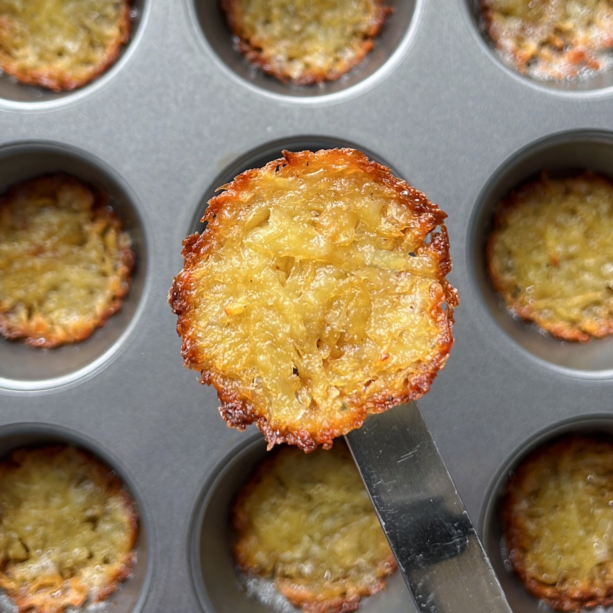 A close-up of a golden-brown, crispy Muffin Pan Potato Rosti held above a muffin tin with similar rosti in each cavity.