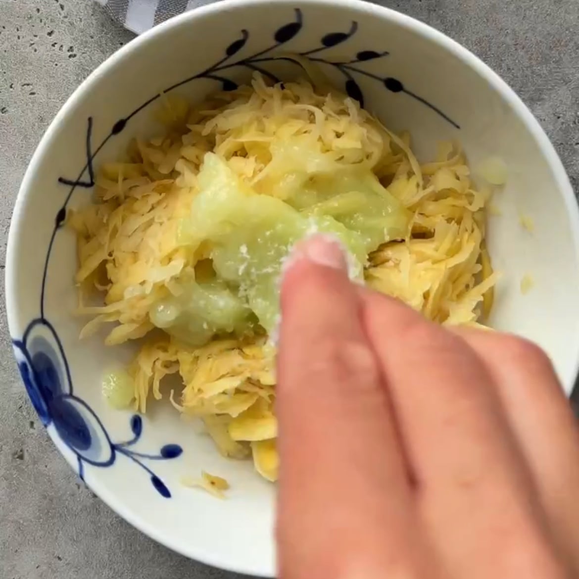 A hand sprinkles salt onto shredded potato and onion in a white bowl with a blue floral design.