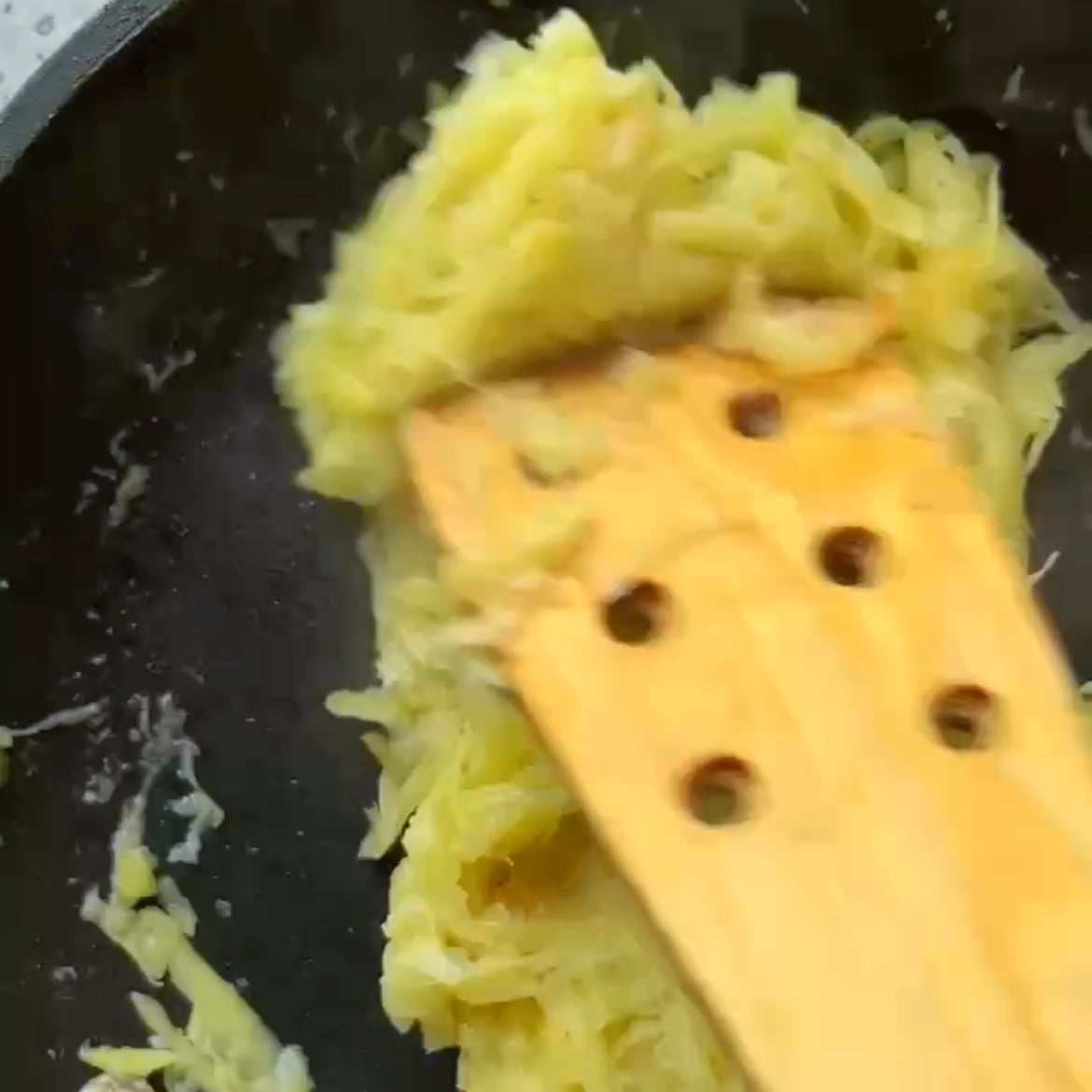 A wooden spatula stirs shredded potatoes and onion in a black frying pan.