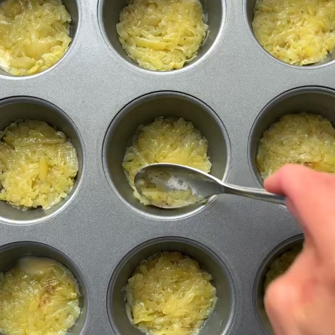 A hand uses a spoon to press shredded potato mixture into a muffin pan.
