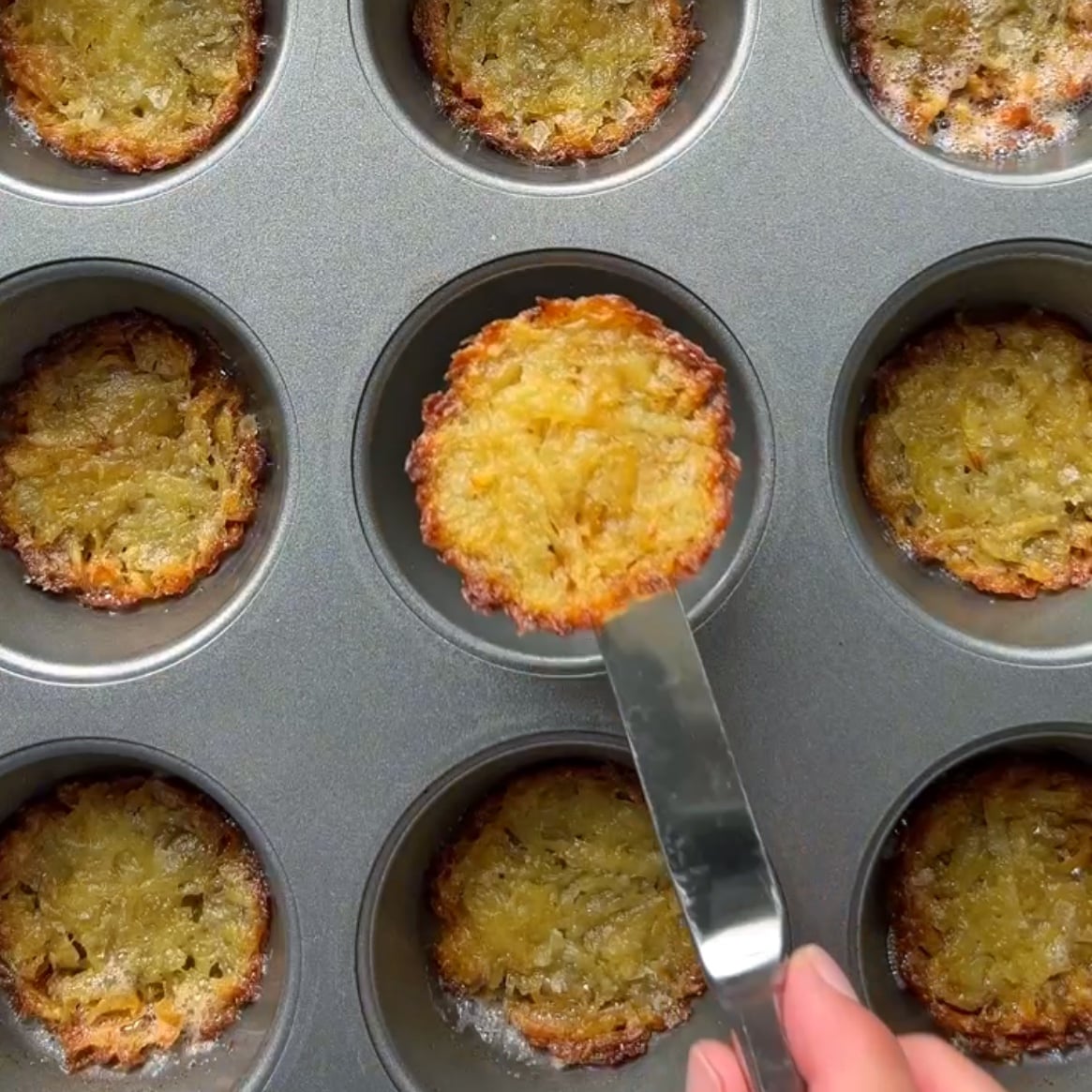 A hand lifts a golden brown Muffin Pan Potato Rosti from a muffin tin filled with similar crispy rosti.