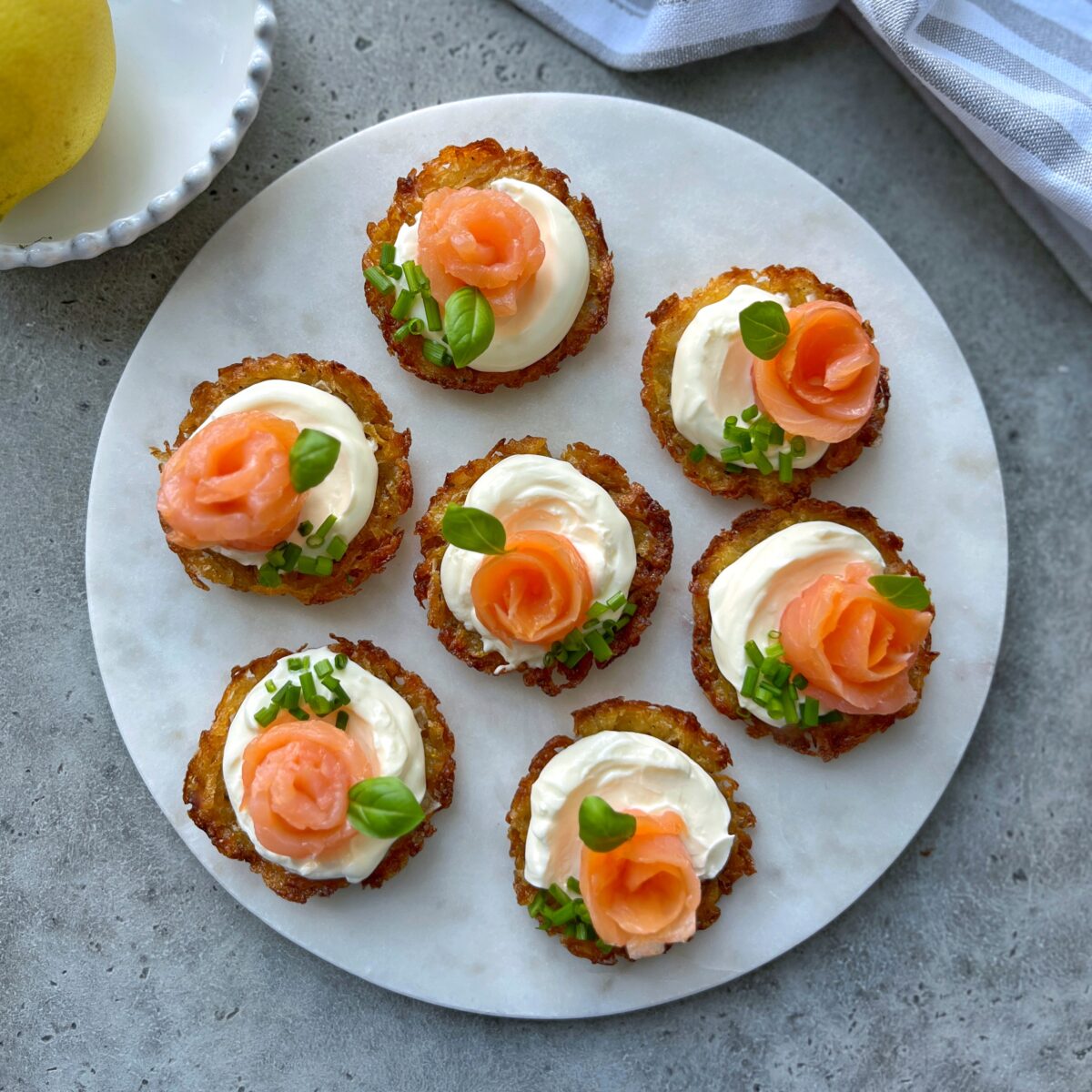 Seven potato bites topped with sour cream, smoked salmon, chives, and basil are arranged on a round white plate set on a gray surface.