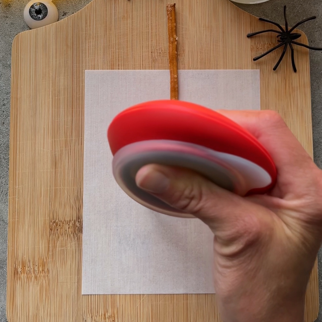 A hand holds a red piping tool over a pretzel stick and a sheet of parchment paper on a cutting board.