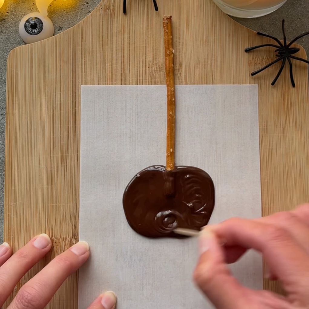 A hand swirls a toothpick in melted chocolate on parchment paper.