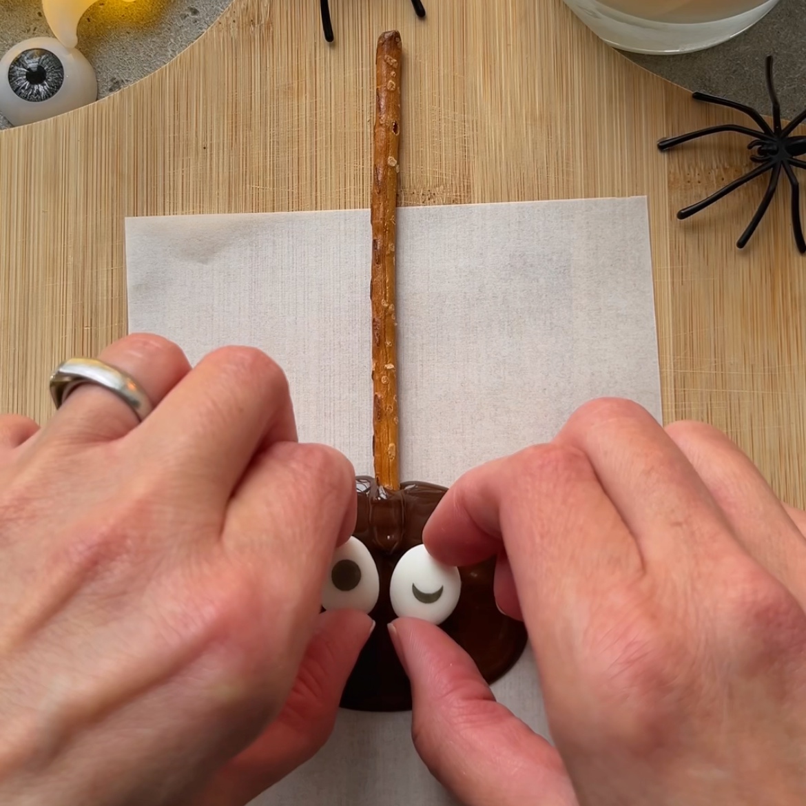 Two hands place candy eyes on a chocolate-covered Pretzel Spiders treat with a pretzel stick handle, on a sheet of parchment paper.