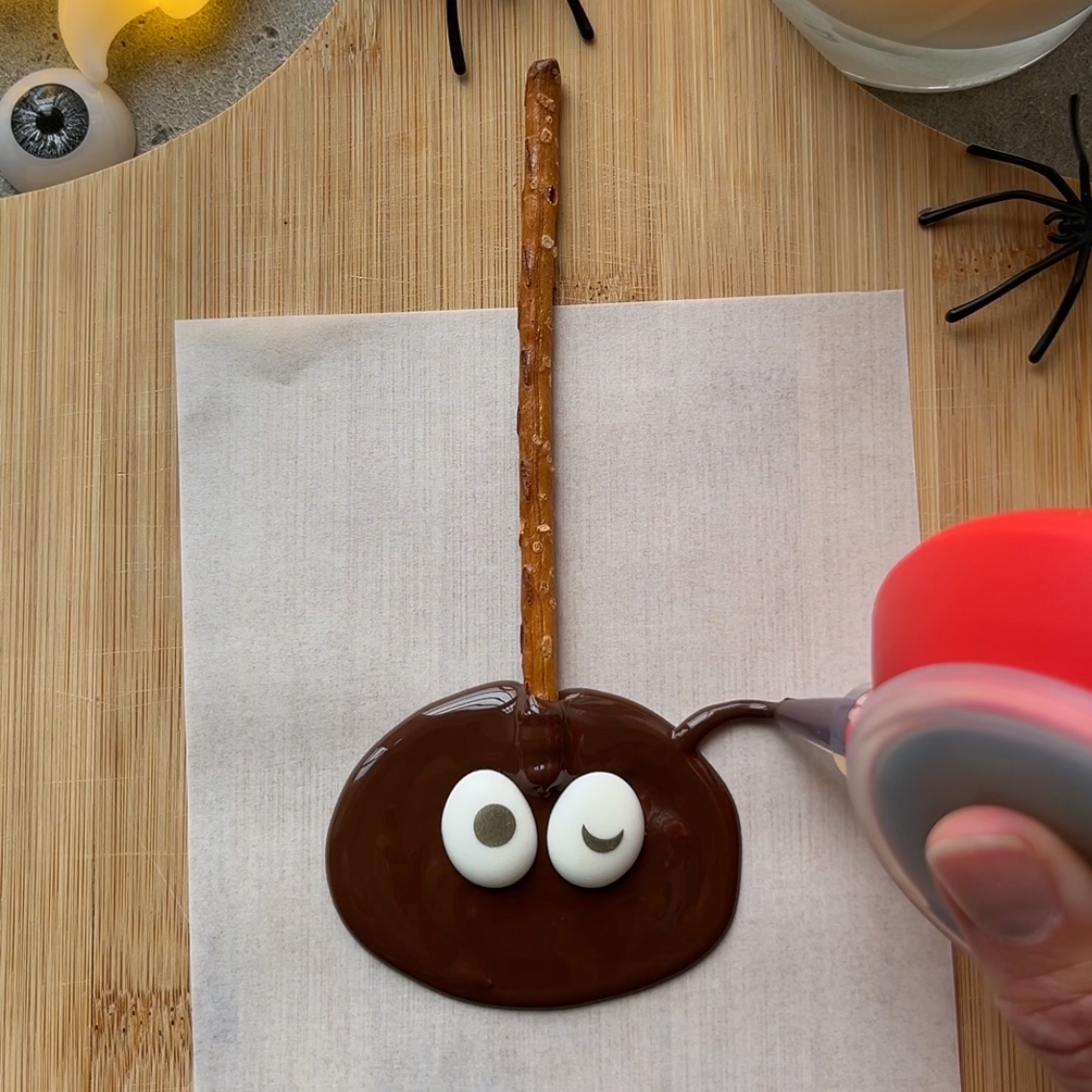 A hand pipes chocolate legs onto a googly-eyed chocolate spider with a pretzel stick handle.