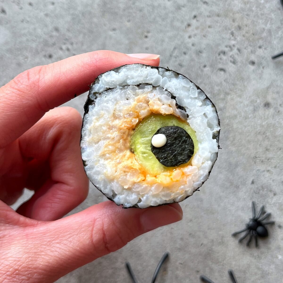 A hand holds a sushi roll designed to look like an eyeball, with rice, seaweed, and other ingredients forming the iris and pupil.
