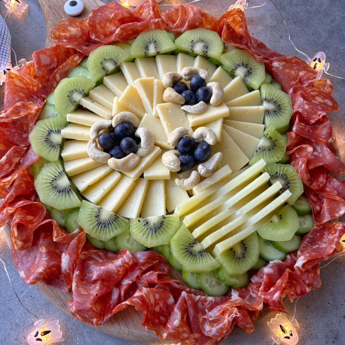A side view of a Spooky Charcuterie Board with sliced cheese, kiwi, cashews, blueberries, and a ring of salami, all arranged in a festive circular pattern.