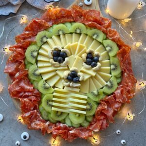 A Spooky Charcuterie Board arranged as a skull with cheese, blueberries, kiwi, cashews, sliced salami border, and surrounded by small ghost string lights.