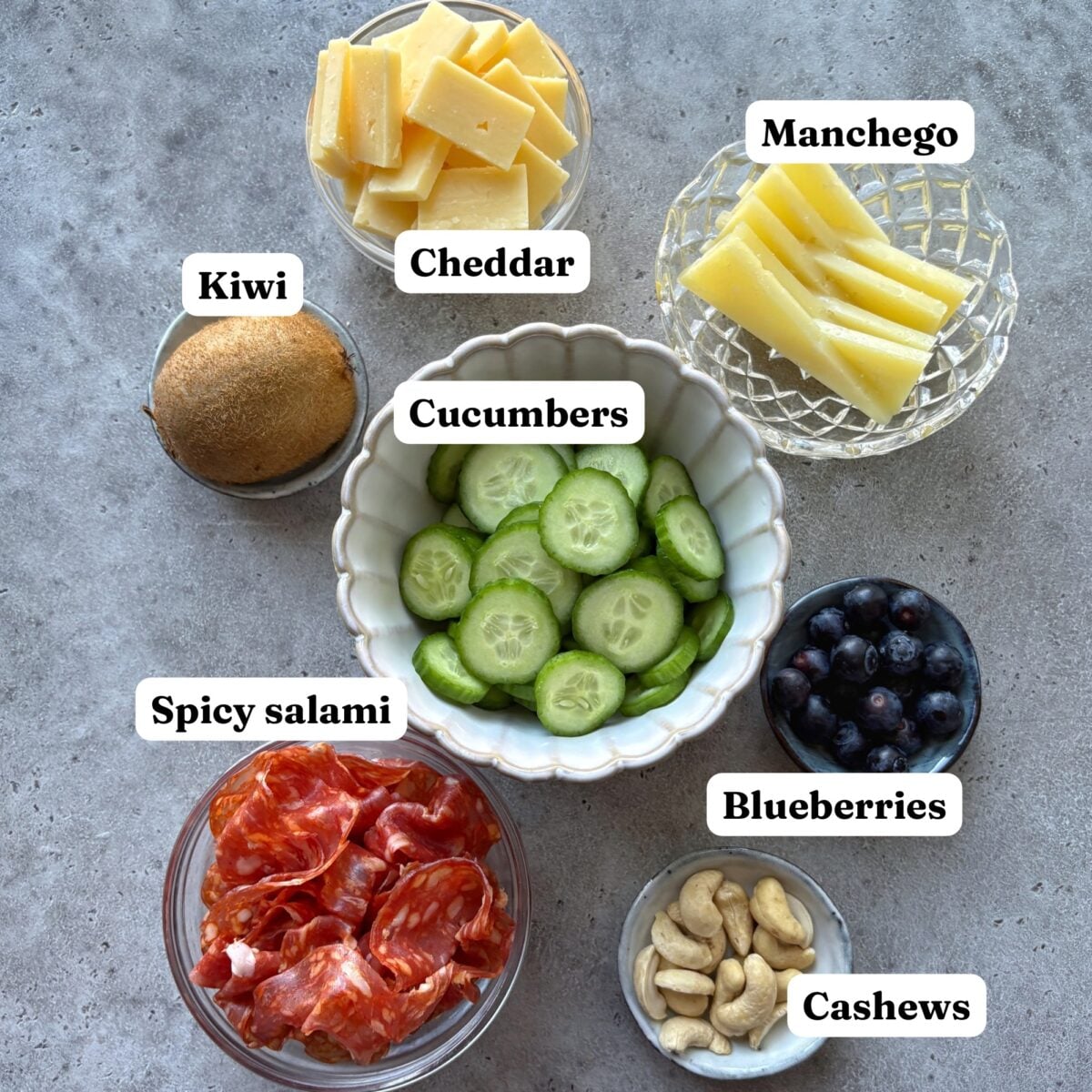 Spooky Charcuterie Board ingredients featuring assorted cheeses, fruits, salami, cucumber slices, blueberries, and cashews arranged in bowls on a gray surface, each labeled.