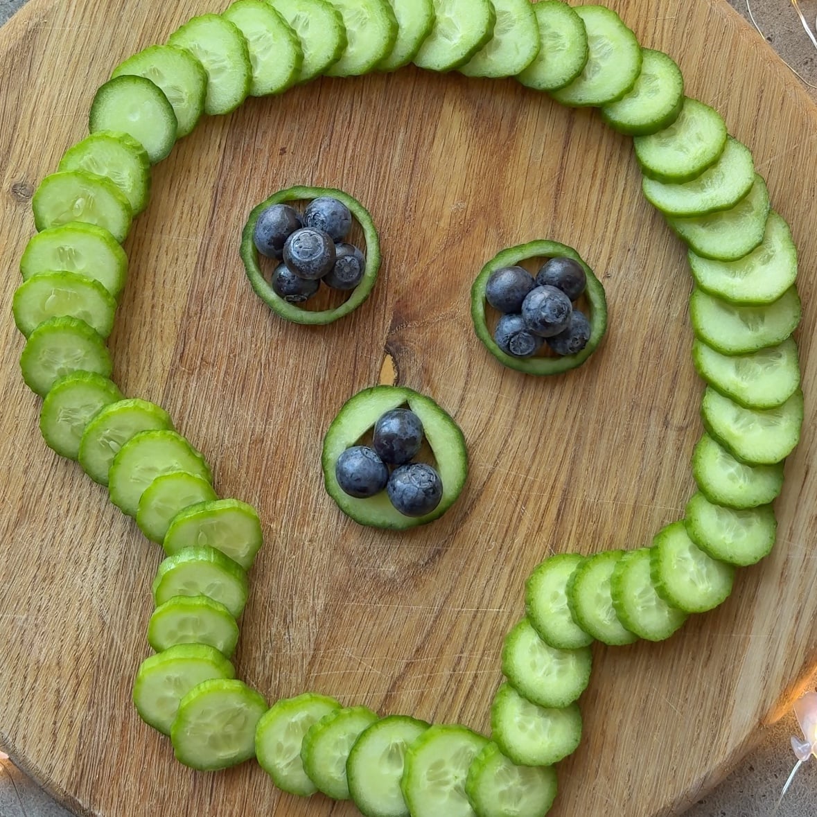 Cucumber slices arranged in the shape of a skull on a wooden board, with blueberries for the eyes and nose.