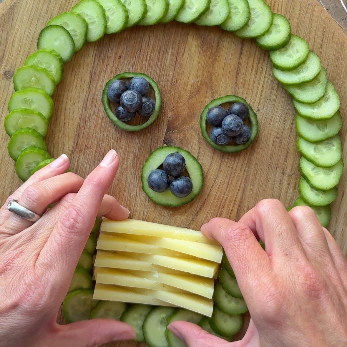 Sliced cucumber and blueberries form a skull on a wooden board, with hands holding stacked cheese slices as a mouth.