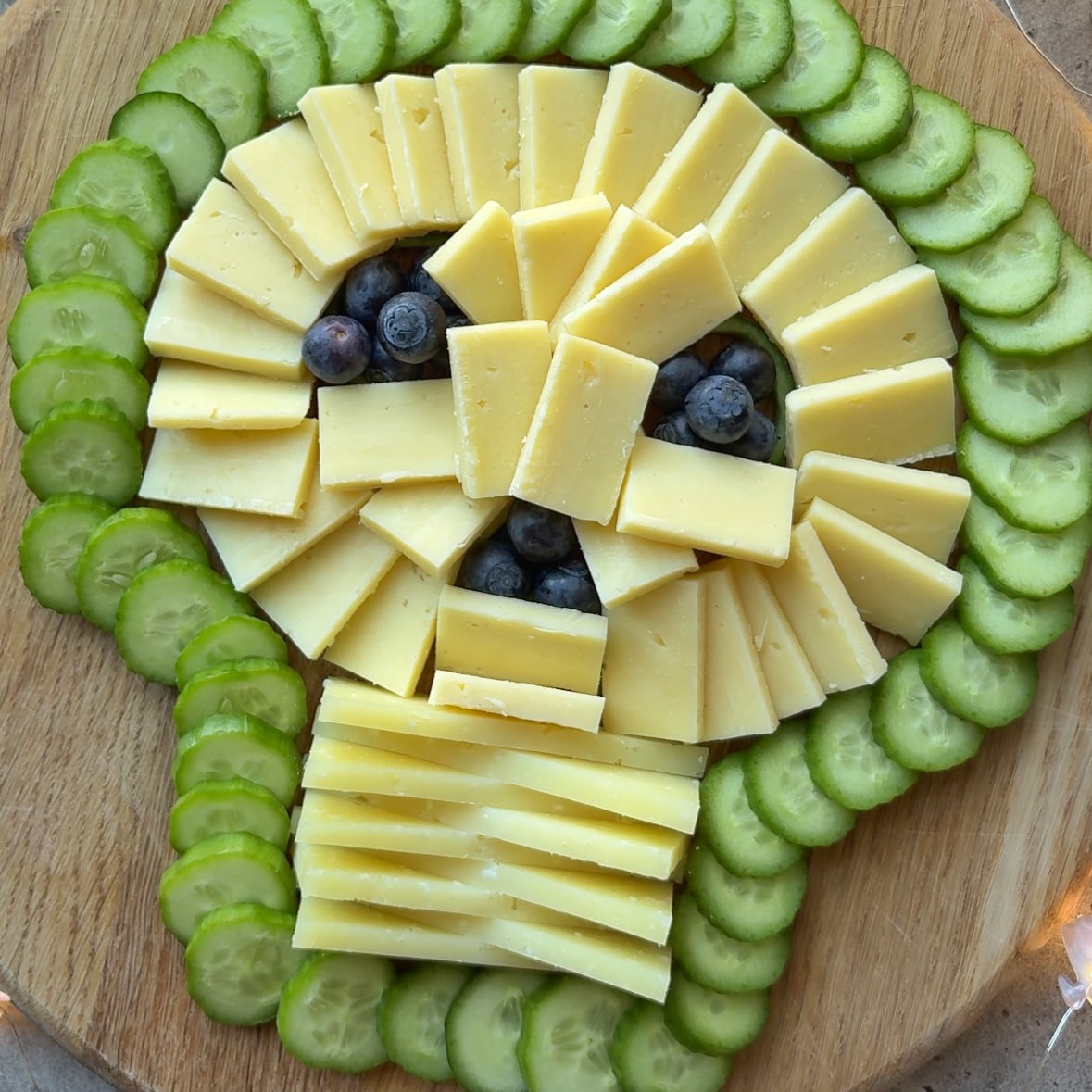 A spooky charcuterie board featuring a cheese and cucumber skull, with blueberries for eyes and nose, arranged on a round wooden board.