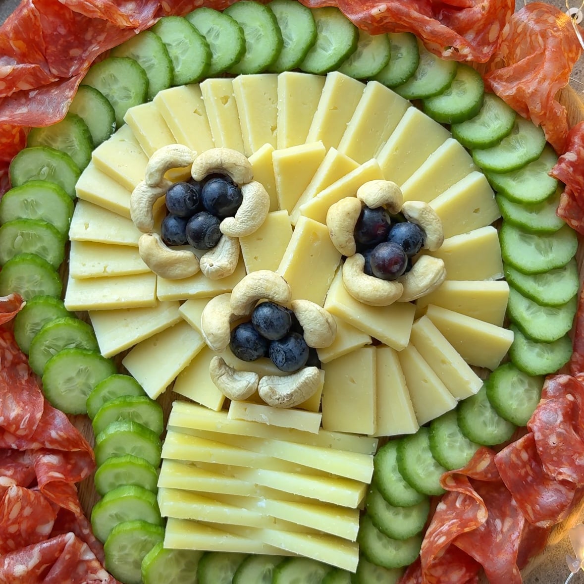 A spooky charcuterie board arranged to resemble a skull, made with cheese slices, cucumber, cashews, blueberries, and salami.