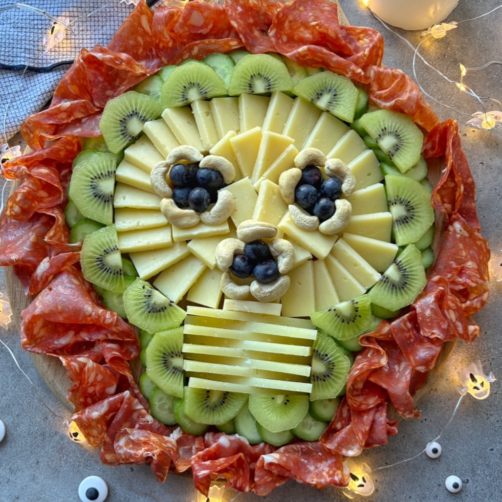 A spooky charcuterie board arranged as a skull with cheese, kiwi, blueberries, cashews, and salami, surrounded by string lights and Halloween-themed decorations.