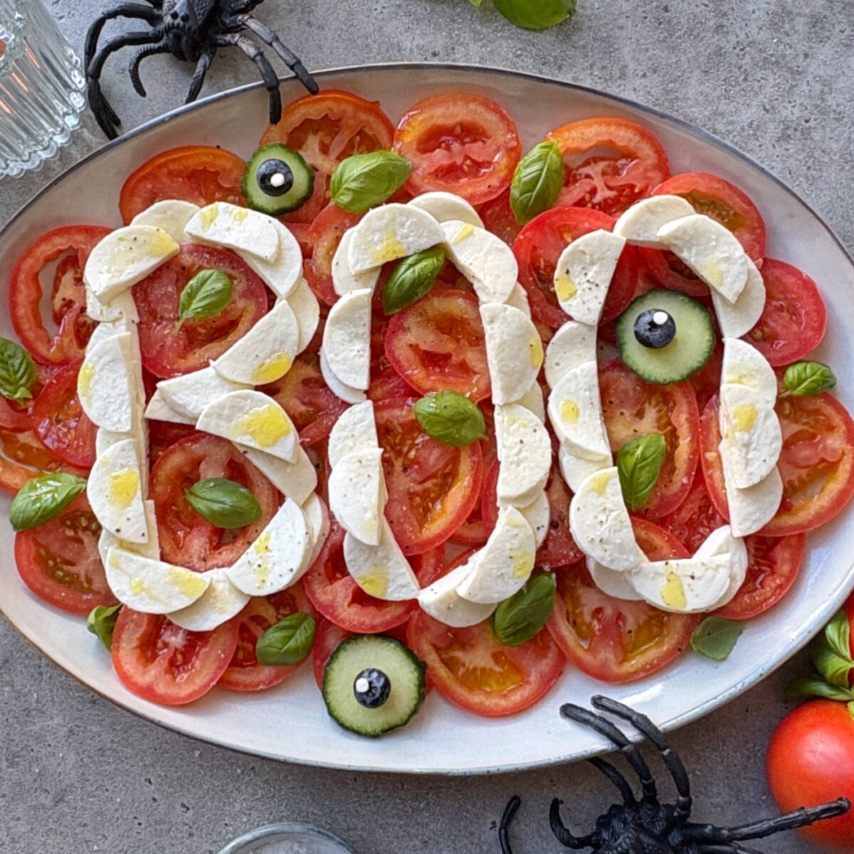 Halloween Salad arranged to spell BOO with mozzarella on sliced tomatoes, garnished with basil, blueberries, and cucumber, placed on a platter with plastic spiders for a festive Halloween theme.